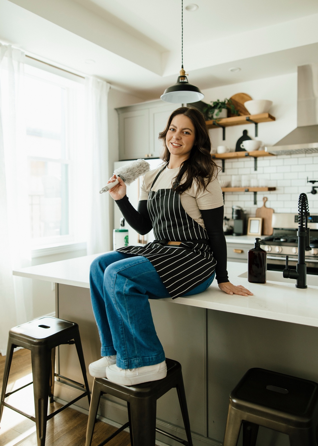 The owner of EVE Home Services sitting on a kitchen island holding a duster.